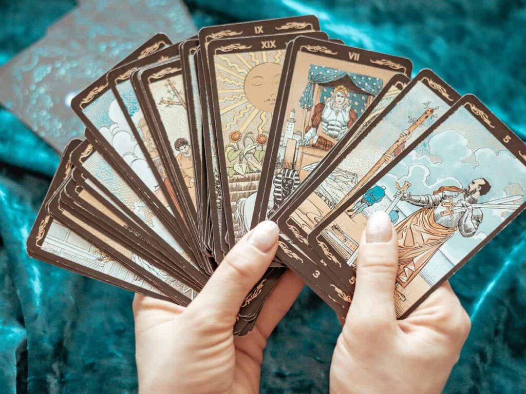 Tarot kartlarını tutan ellerin yakın plan görüntüsü, destenin üzerinde çeşitli sembollerin ve tarot karakterlerinin bulunduğu detaylar yer alıyor. Arka planda ise gizemli bir atmosferi destekleyen kadife benzeri bir yüzey var.