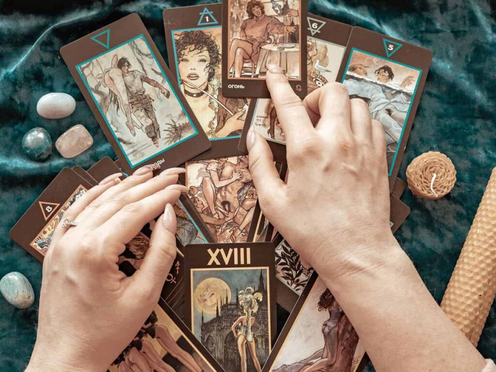 Elleriyle tarot kartlarını tutan bir kişinin tarot kart açılımı yaparken görüldüğü, taşların ve mumların yer aldığı bir fal sahnesi. Kartların üstünde çizgi roman tarzı illüstrasyonlar yer alıyor.