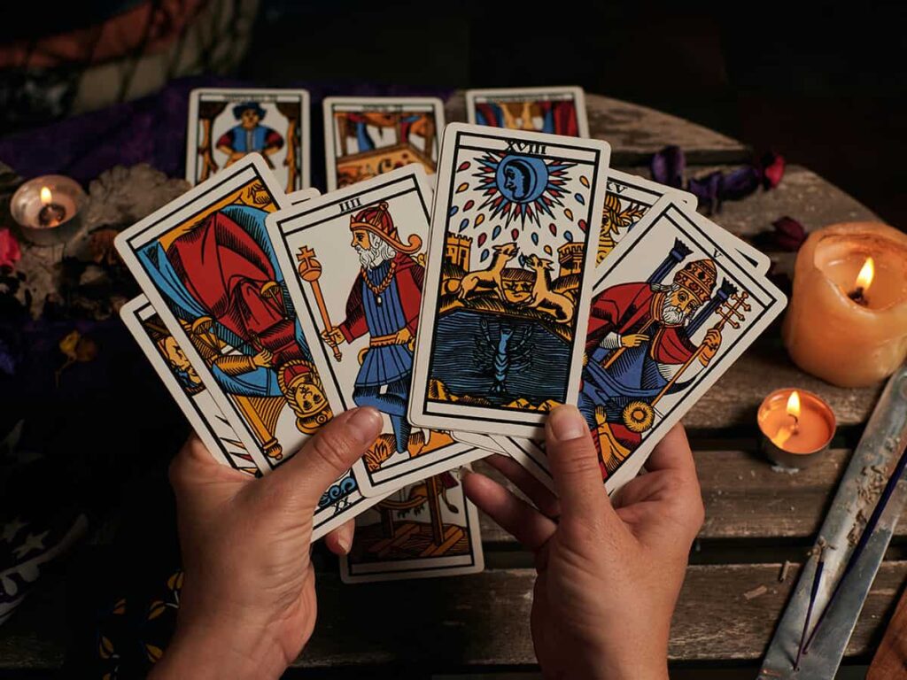 Bu fotoğraf, Tarot destesinde bulunan "Ay" kartını gösteriyor. Kartın ortasında büyük bir dolunay, etrafında yıldızlar ve denizin üzerinde yansıması var. İki köpek veya kurt, ayın karşısında havluyor. Ayın altında suya doğru çıkan bir yengeç figürü yer alıyor. Kartın genel havası gizemli ve karanlık. Bu kart, sezgileri, bilinçaltını ve gizemleri temsil eder.